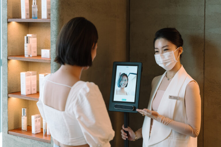 Woman receiving digital skin analysis from a masked beauty consultant in a modern clinic with product shelves.